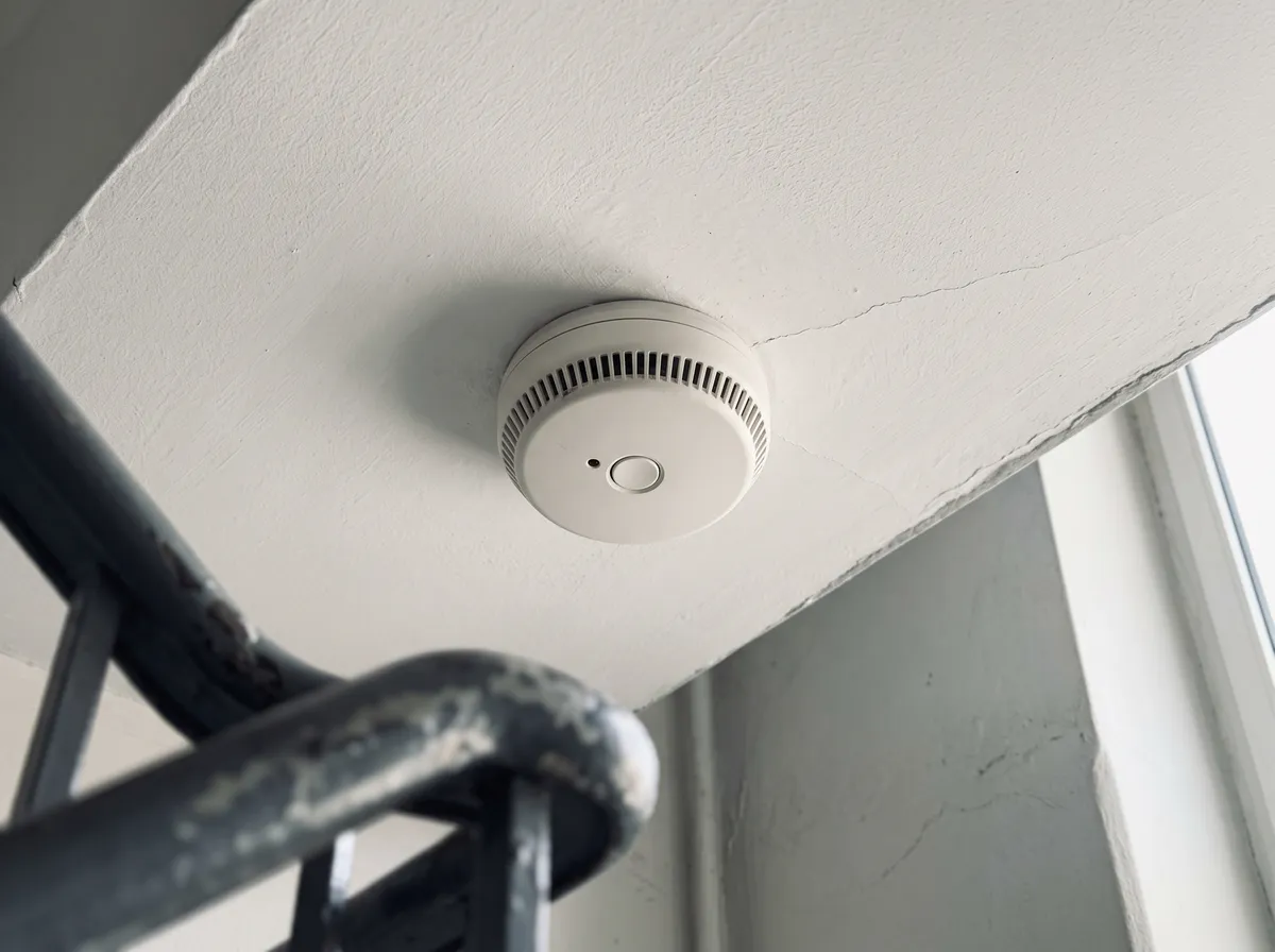 Weißer Rauchwarnmelder an der Decke eines Treppenhauses, davor ein Treppengeländer
